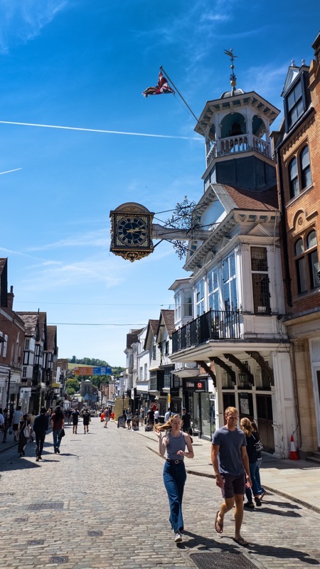 Shoppers on the cobbled slope of Guildford High Street beneath the projecting gilded Guildhall clock